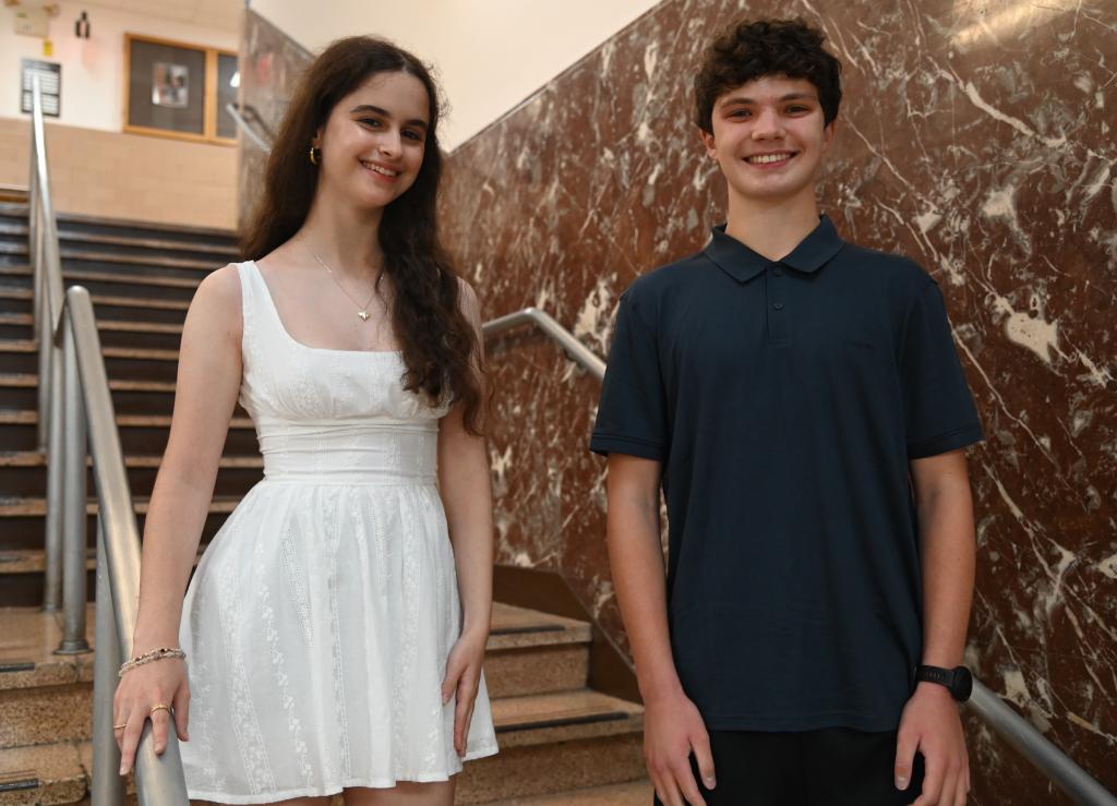 A young woman in a white dress and a young man in a dark polo shirt stand smiling near a staircase.